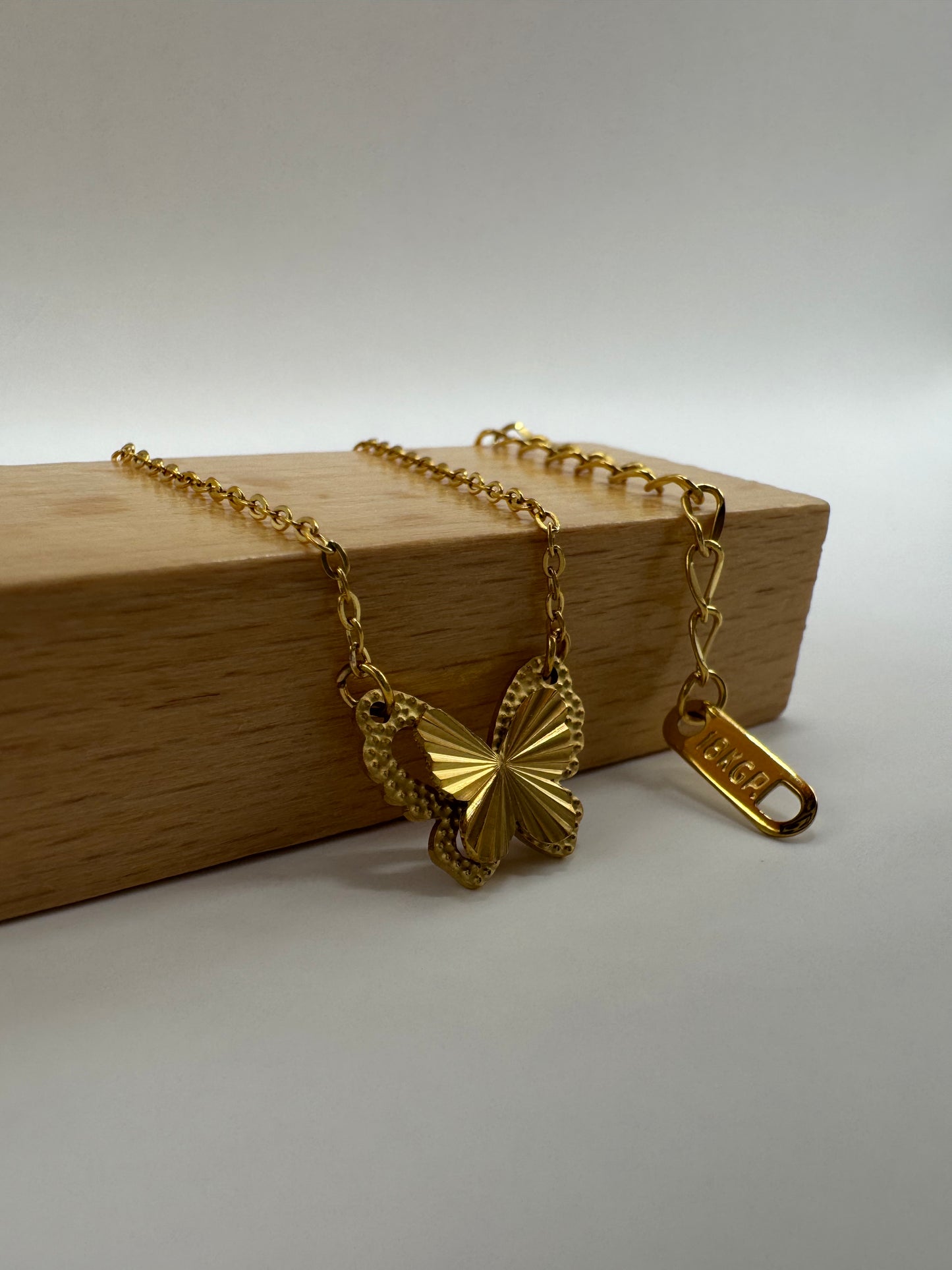 Close-up Aurora Clásica gold butterfly necklace displayed on wooden block with chain and clasp visible.