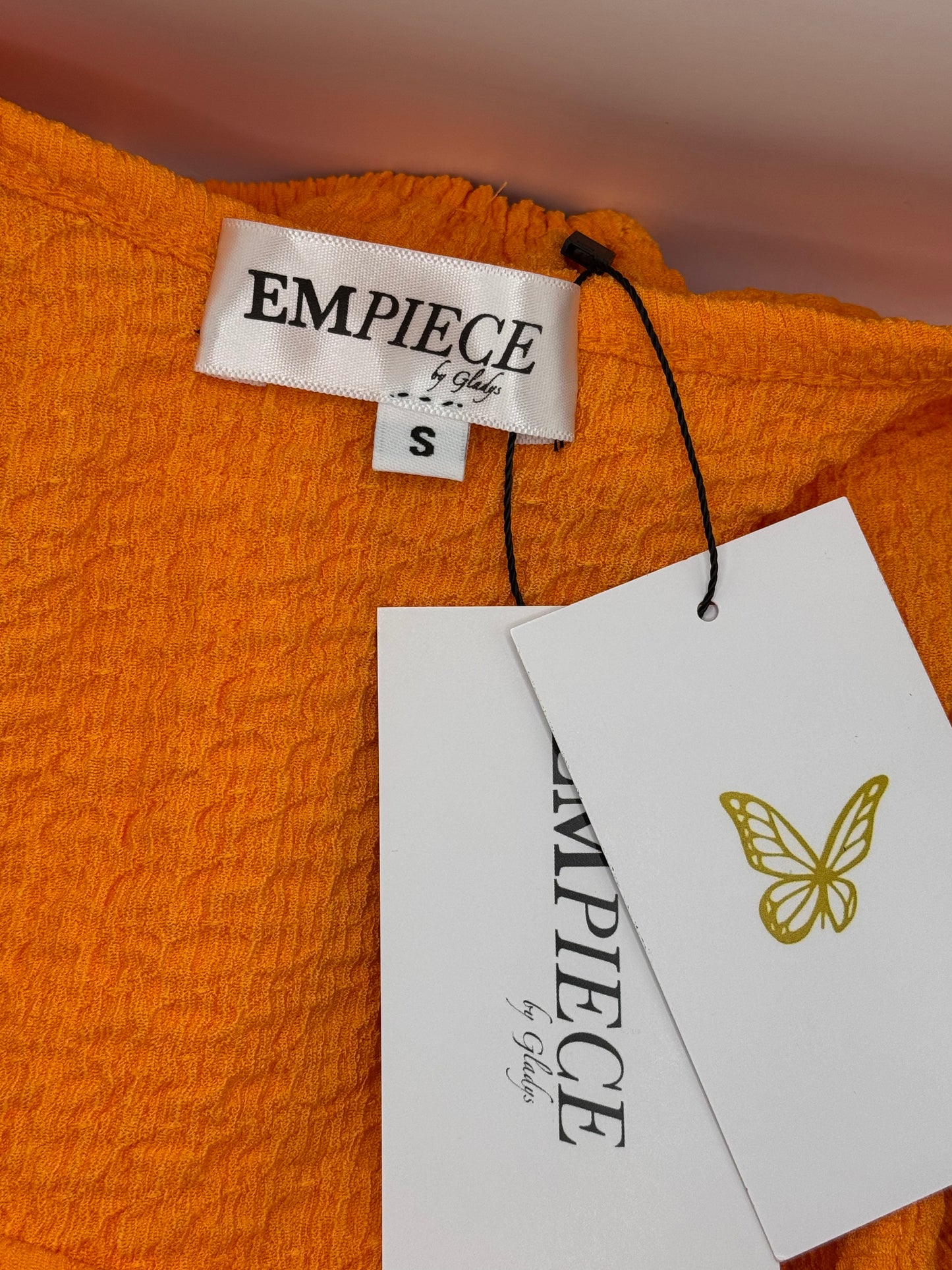 Close-up of EMPIECE™ by Gladys woven label and butterfly hang tag attached to The Scarlet Allure Top in orange fabric.