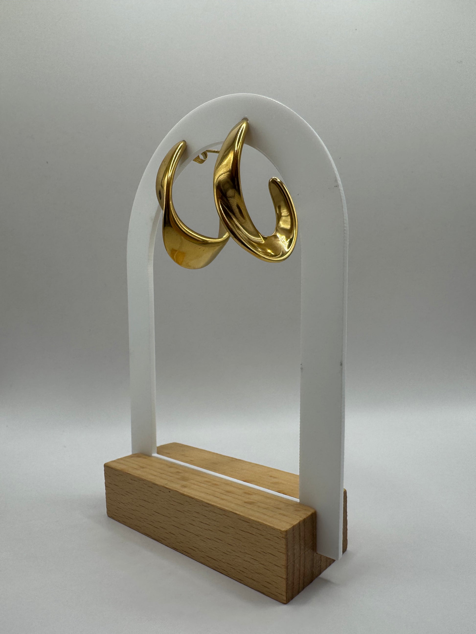 Gold sculptural C-shaped hoop stud earrings displayed on clear acrylic stand with wooden base, The Virelle Earrings by EMPIECE™.
